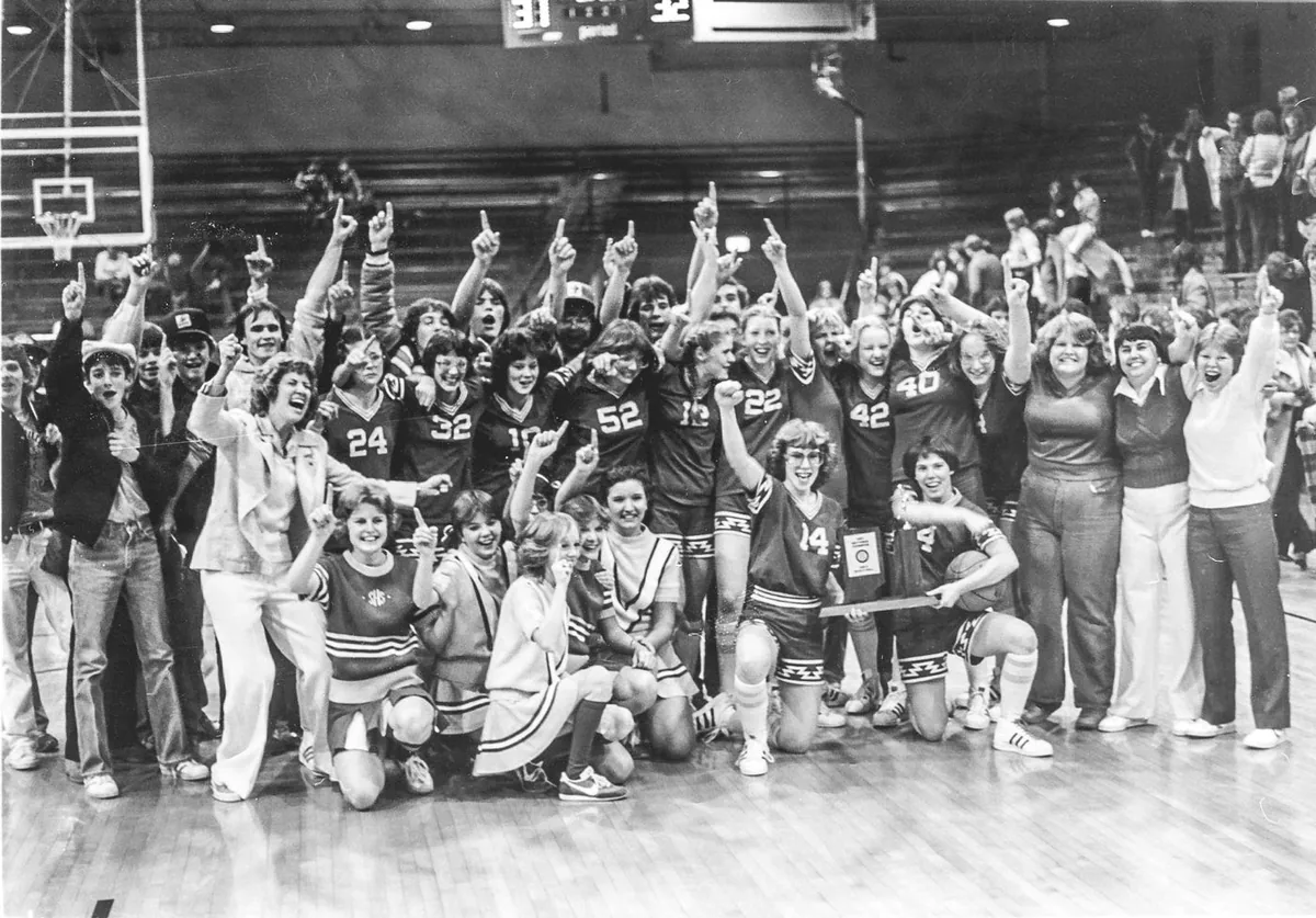 The Warriorettes, cheerleaders and fans crowd together on the floor holding up index fingers, a trophy plaque in front, the scoreboard above reading 31 and 32