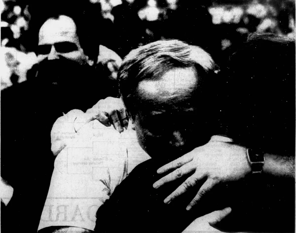 Newspaper photo: Austin's coach, eyes shut, in a long embrace, another man's hand on his back, a crowd behind