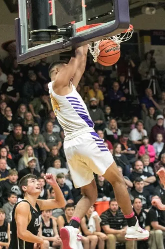 A Scottsburg player in white hangs on the rim after a dunk, an Austin player below, the stands full behind