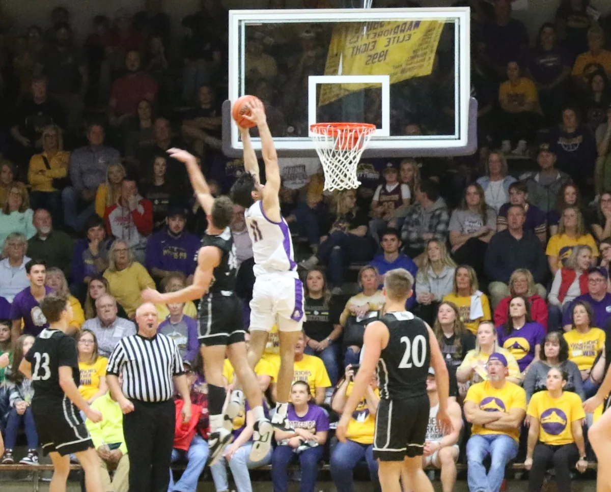 A Scottsburg player in white rises for a two-handed dunk over an Austin defender in black, a referee and a full crowd in purple and gold behind