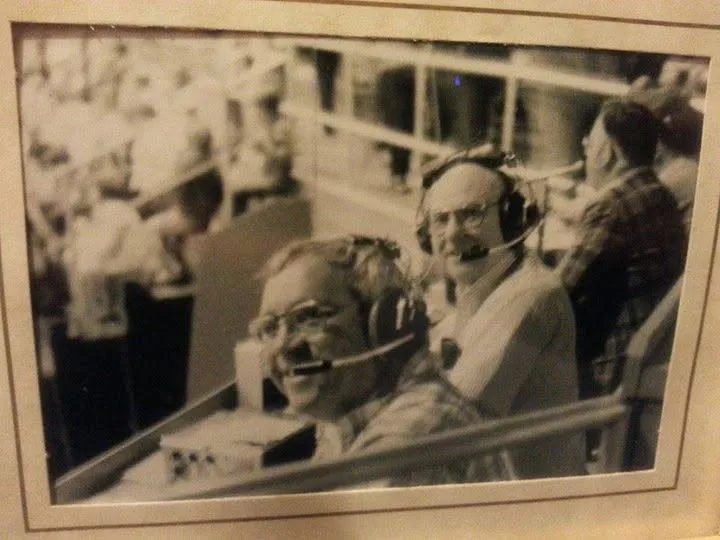 Two men in headsets and microphones smiling at the camera from the broadcast table above a packed crowd
